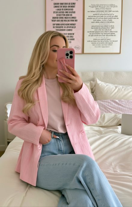 Woman in a pink blazer and jeans sitting on a bed, taking a mirror photo